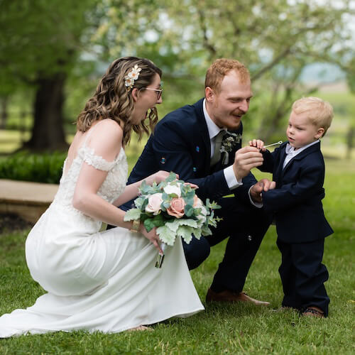 Happy groom and bride blowing bubbles with ring bearer in scenic pasture.