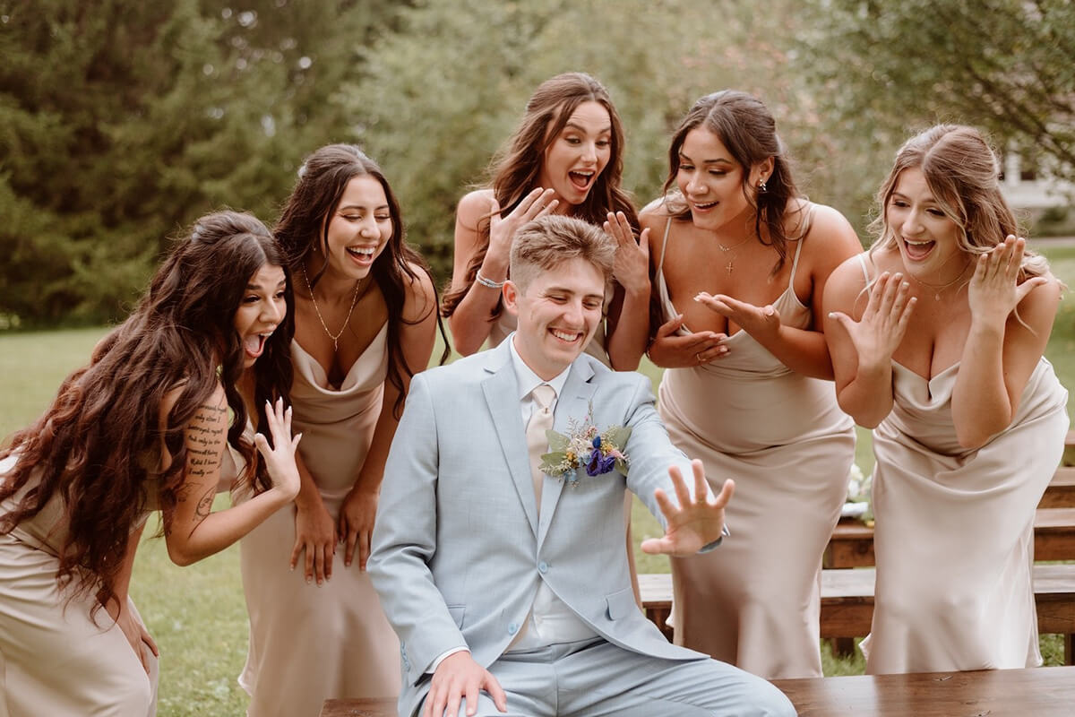 Bridesmaids gasping with joy surrounding newly married groom looking at his wedding band.
