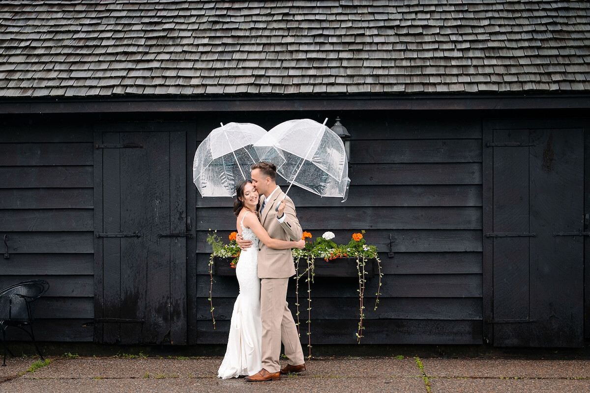 Happy groom and bride holding umbrellas in front of rustic barn building.