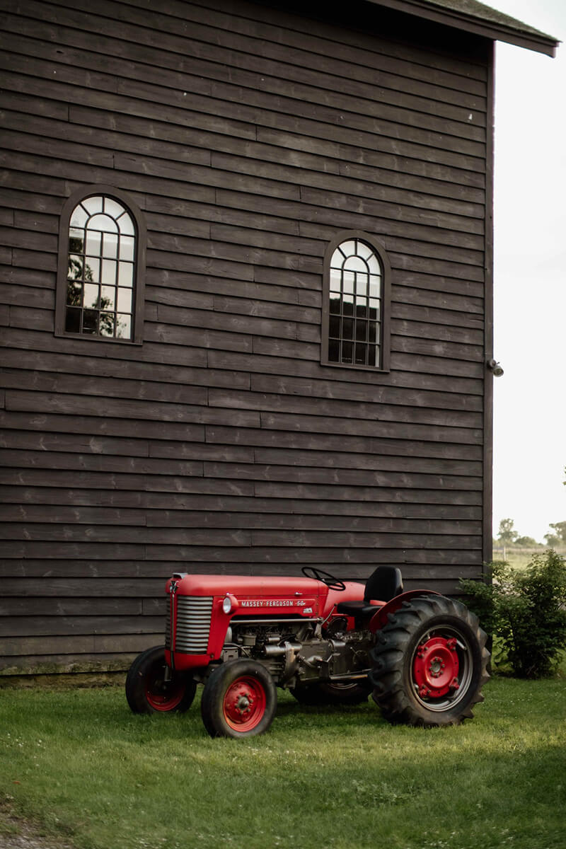Red tractor parked outside brown rustic barn with two windows.