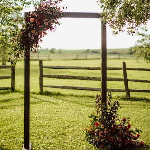 Square chuppah decorated with flowers for ceremony in front of scenic pasture.