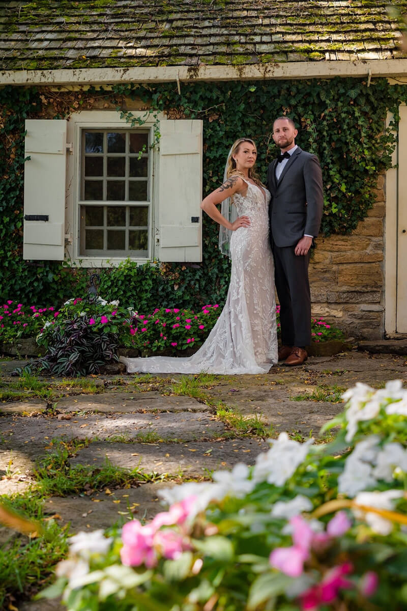 Married couple posing outside of brick house surrounded by pink and white flowers.
